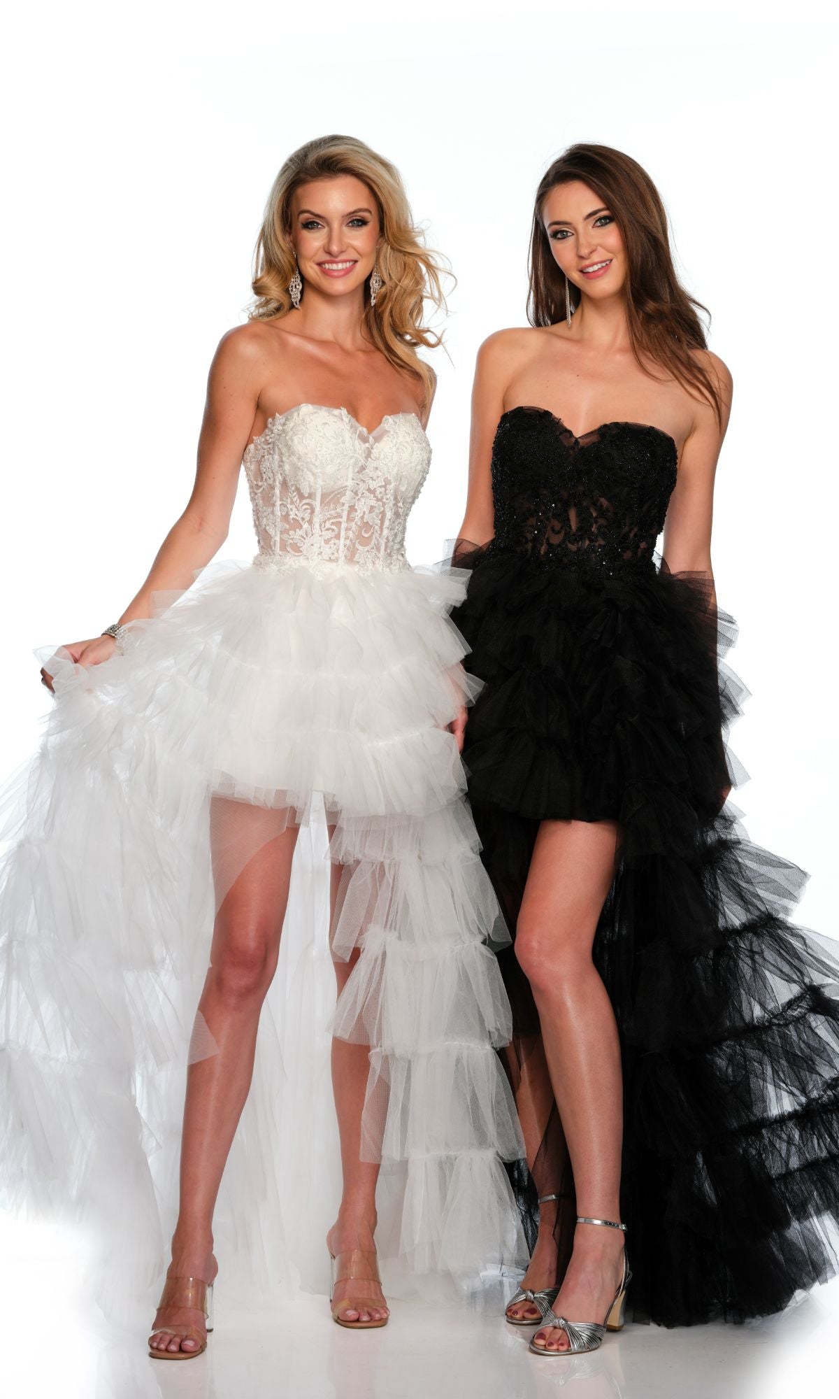Two women stand and smile against a white background, each wearing the Long Formal Dress 11651 by Dave and Johnny—strapless with a lace corset bodice and ruffle layered tulle skirt, one in white and the other in black.